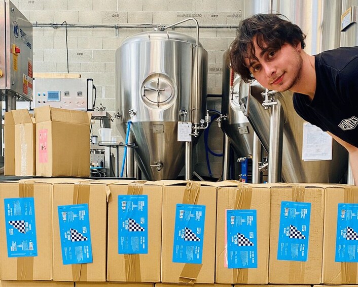 A person in a black shirt leaning over a row of cardboard boxes with blue labels, in a room with stainless steel brewing equipment. 3