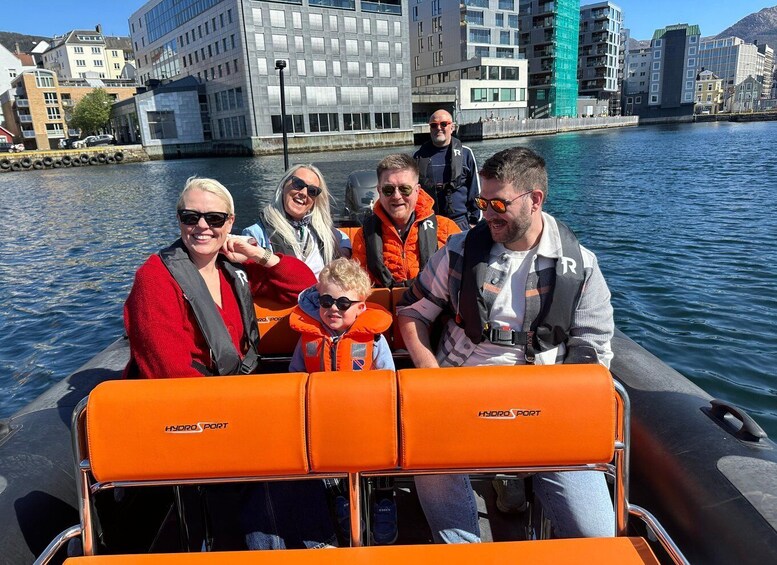 A group of people on a boat with orange seats labeled 'HYDROSPORT' in a city setting with buildings and a body of water. 2