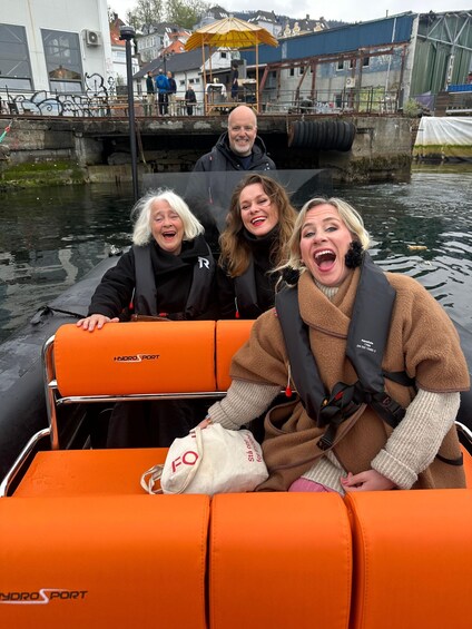 Three people on an orange inflatable boat with 'Hydroport' branding, on a body of water with industrial buildings in the background. 3