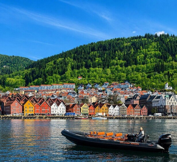 A boat named 'DISCOVER BERGEN' on calm water with a colorful town and forested hills in the background. 1