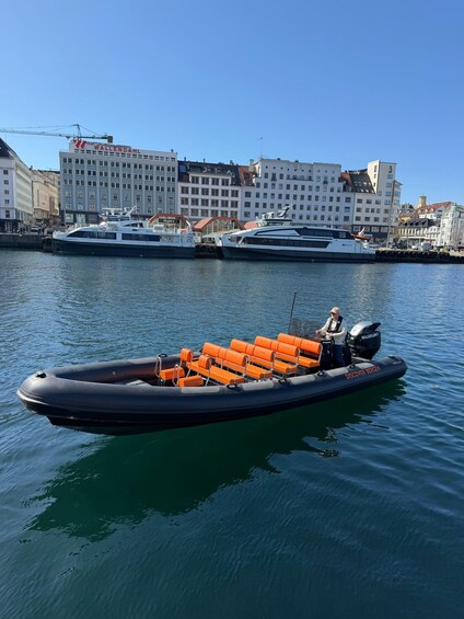 A small inflatable boat with orange seats docked near a waterfront with buildings in the background. 1