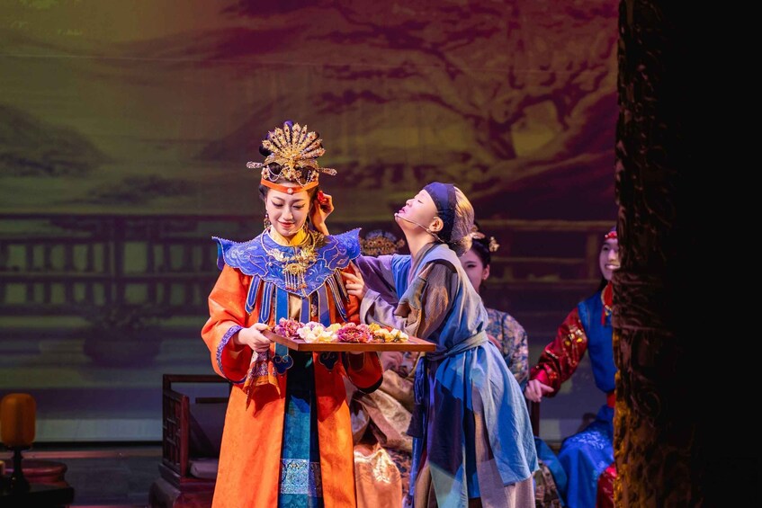A person in traditional attire holding a tray with flowers, standing in front of a backdrop with a bridge and trees. 4