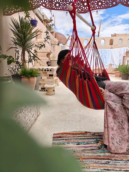 A person relaxing in a colorful hammock outdoors, surrounded by potted plants and a traditional building. 1