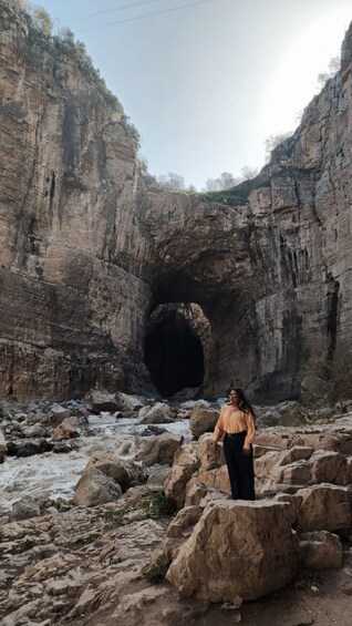 A person standing on rocks near a cave entrance with a river flowing through a rocky canyon. 3