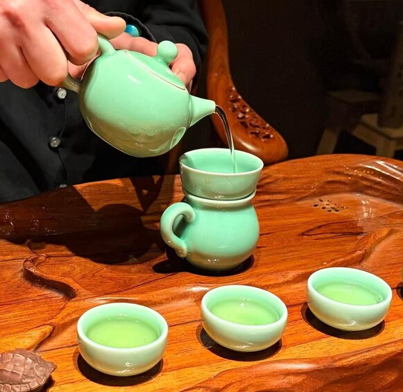 A person pouring tea from a teapot into a cup, with several cups on a wooden tray. 1