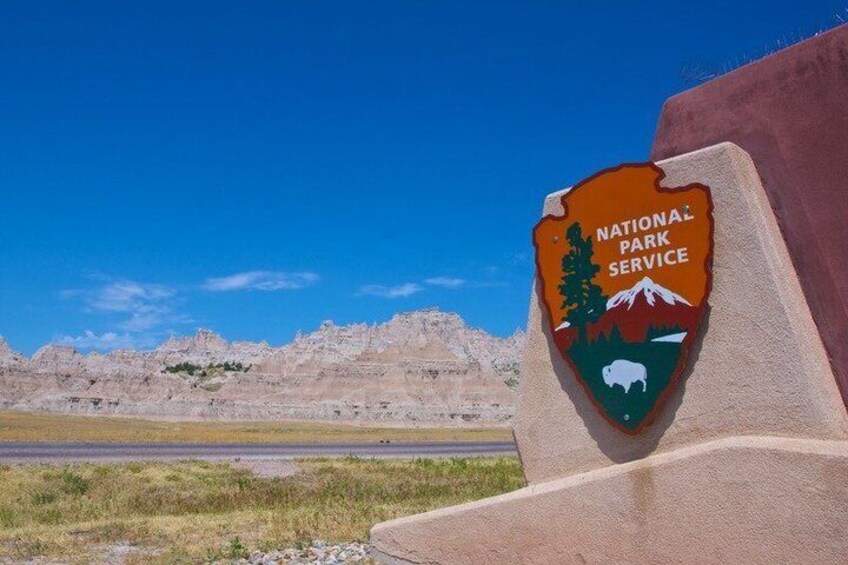 A National Park Service sign with a mountain and bison graphic, set against a backdrop of a clear blue sky and a rocky landscape. 5