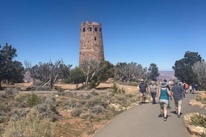 A group of people walking on a path towards a tall, ancient tower in a dry, open landscape with sparse vegetation. 1
