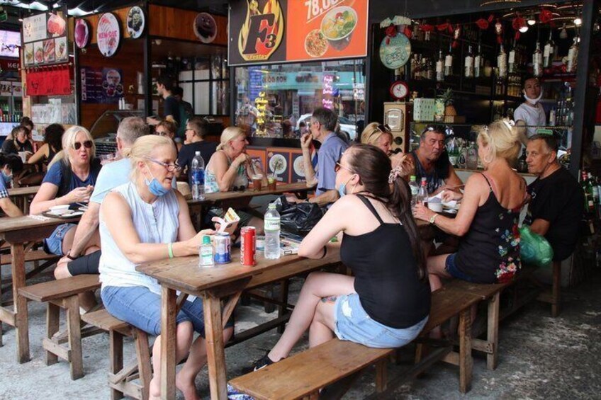 People sitting at wooden tables in an outdoor cafe with food and drink options visible. 4