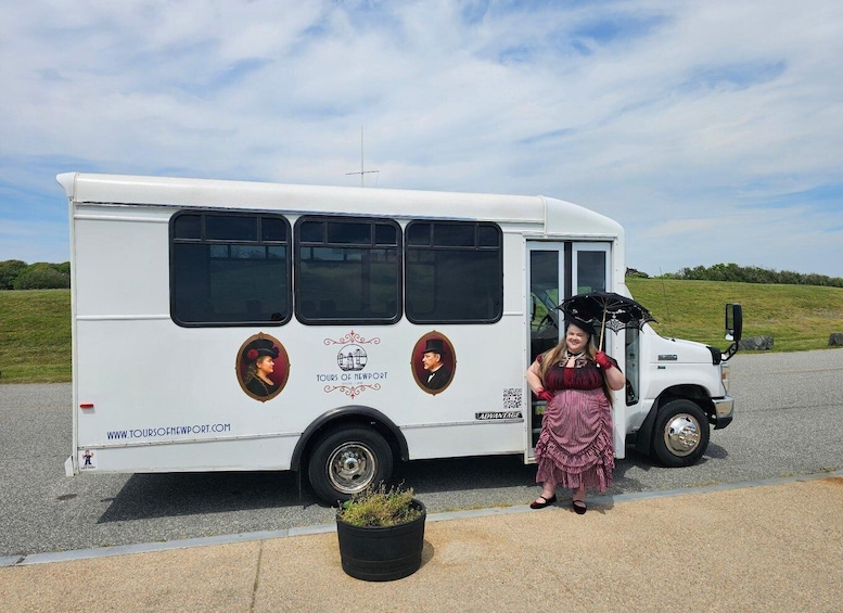 A white bus with 'TOURS OF NEWPORT' and 'WWW.TOURSOFNEWPORT.COM' on its side, parked on a road with a grassy hill in the background. 1