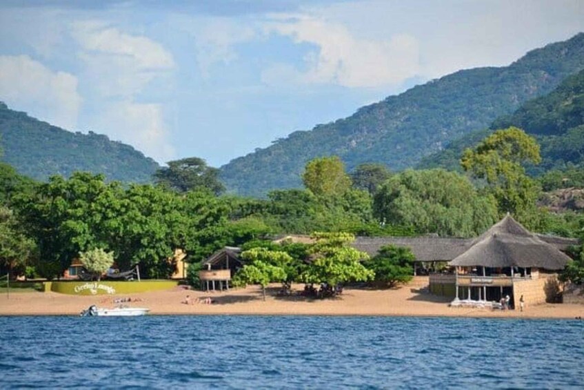 A beach with thatched-roof huts, a boat, and lush green hills in the background. 1