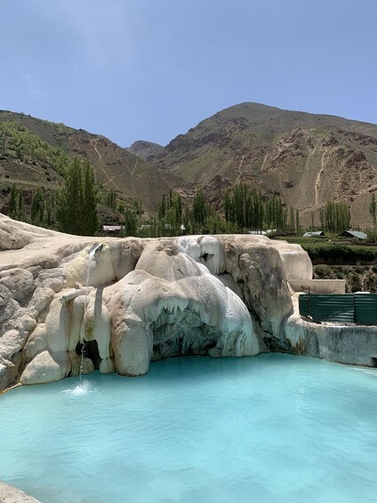 A turquoise hot spring with white travertine formations, surrounded by mountains and sparse vegetation. 5