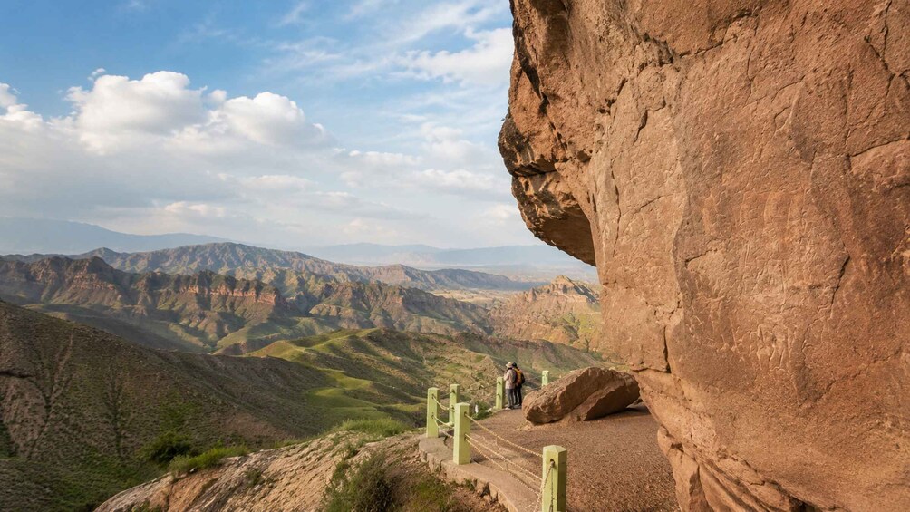 A rocky cliff with a person standing on a ledge, overlooking a valley with mountains in the distance. 3