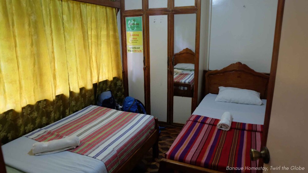 A room with two beds, one with a striped bedspread and the other with a plain one. There's a blue bag on the floor and a check-out sign on the wall. 5