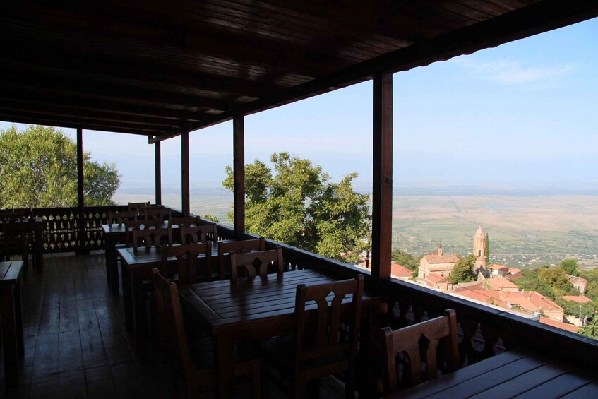 An outdoor dining area with wooden tables and chairs, overlooking a valley with buildings and a church. 2
