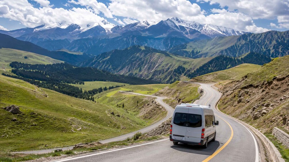 A white van driving on a winding mountain road with snow-capped peaks in the background. 4