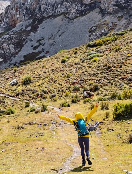 A hiker with arms raised, wearing a yellow jacket and blue backpack, walking on a grassy trail with rocky mountains in the background. 2