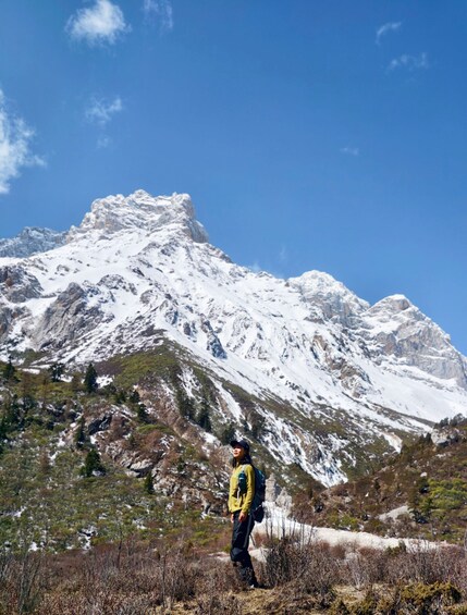 A hiker in a yellow jacket and black pants standing in a mountainous area with snow-covered peaks under a clear blue sky. 3