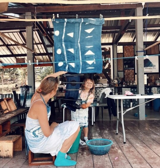 A woman and a child in a rustic outdoor setting, possibly a market or workshop, with a blue patterned cloth hanging above them. 2