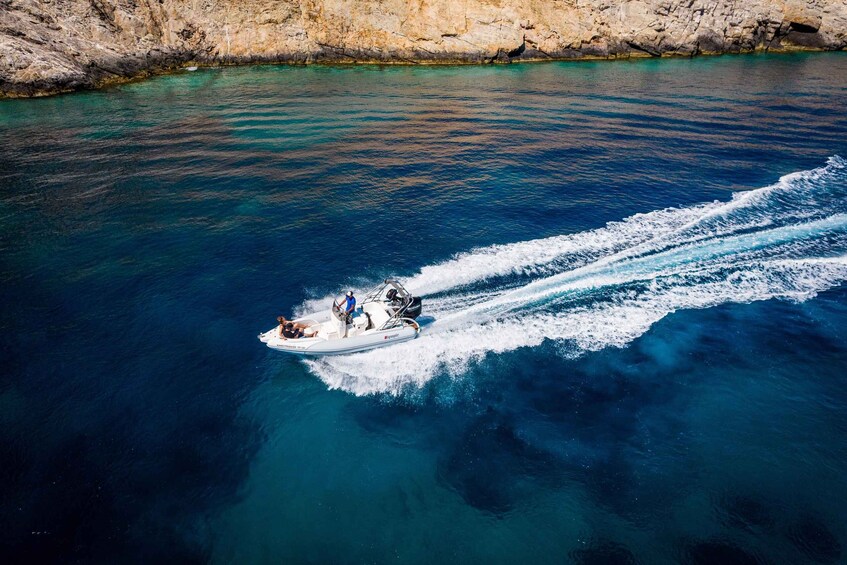 A speedboat with passengers moving through clear blue water near a rocky coastline. 4