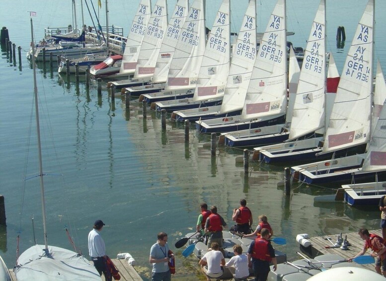 A marina with sailboats docked in a row, people near the boats, and a calm water surface. 3
