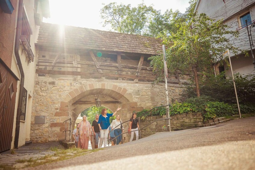A stone archway with people standing beneath it, a street, and a building with a sloped roof. 3