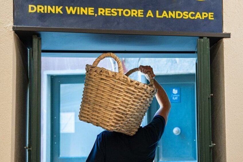 A person carrying a woven basket over their head, with a sign above that reads 'DRINK WINE, RESTORE A LANDSCAPE.' 2