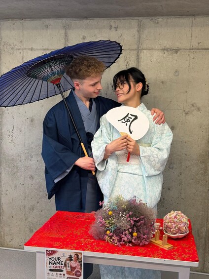 A man and a woman standing in front of a table with a red cloth, a decorative flower arrangement, and a sign with a name in Kanji. 5