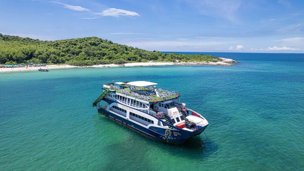 A boat with a deck area and a lifeboat on a turquoise sea, surrounded by a beach and forested land. 1