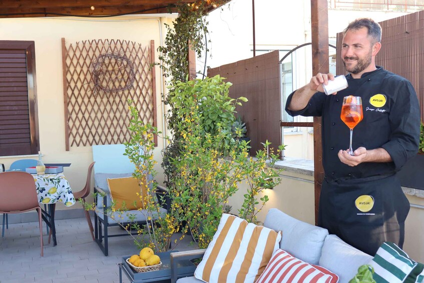 A person in a chef's uniform holding a glass with a drink, standing in an outdoor patio area with a table, chairs, and plants. 5