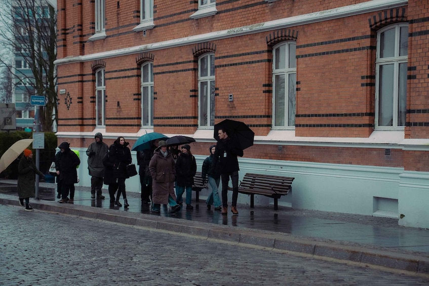 A group of people walking on a wet pavement in front of a red brick building with white trim and multiple windows. 3