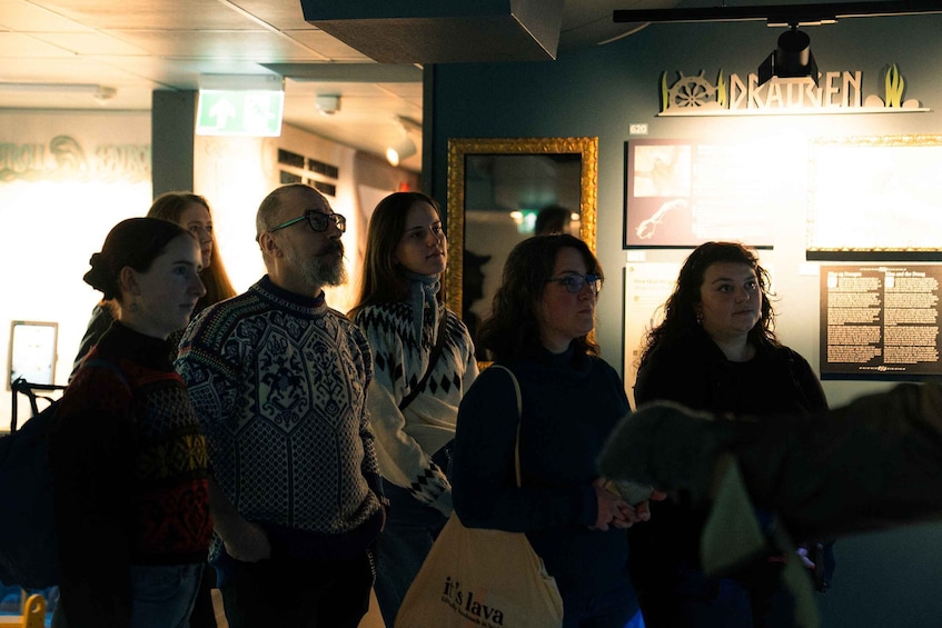 A group of people standing indoors, possibly in a museum or gallery, with a sign that reads 'DRATREN' and a framed picture on the wall. 5