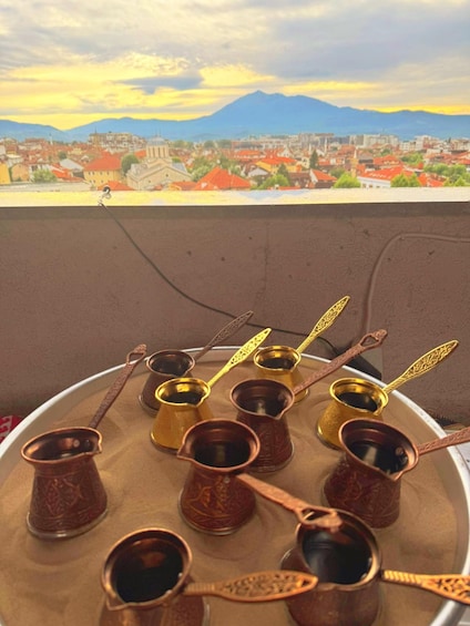 A tray of traditional copper cups with ornate handles, set against a backdrop of a cityscape with mountains. 1