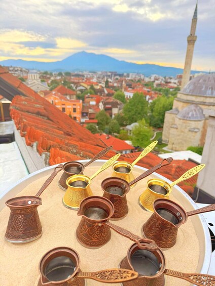 A tray of traditional copper tea cups with ornate handles, set against a cityscape with red-roofed buildings and a mosque with a minaret. 2