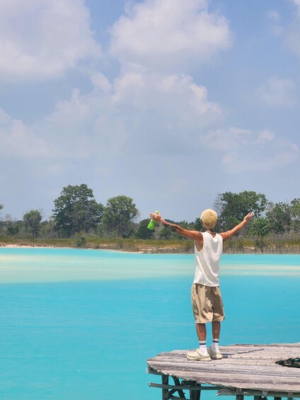 A person standing on a wooden dock by a turquoise sea, holding a green bottle. 4