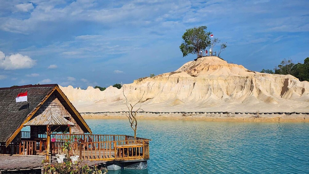 A wooden structure with a flag, a clear blue lake, and a sandy hill with trees. 3