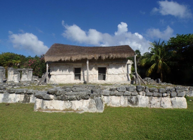 A thatched-roof structure with stone walls, surrounded by a stone perimeter, set in a grassy area with palm trees in the background. 5