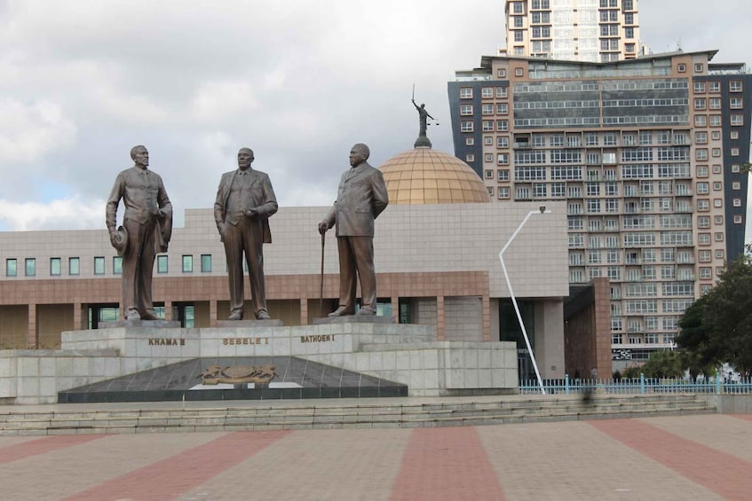 A statue of three men with inscriptions, a dome, and a modern building in the background. 2