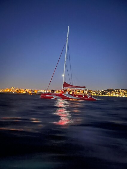 A sailboat with a red and white hull named 'BLUE VISION' on calm water with city lights reflecting on the surface. 2
