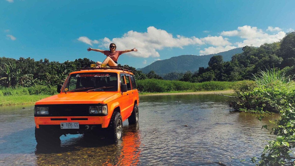 A person standing on an orange vehicle in a shallow river surrounded by greenery and hills. 3