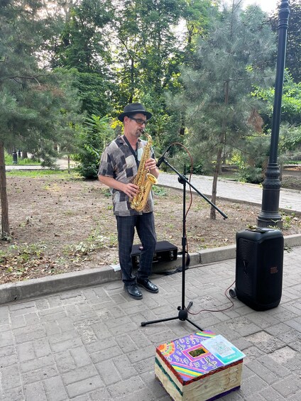A man playing a saxophone in a park with trees and a microphone stand. 5