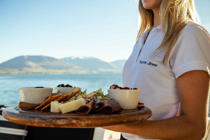 A person holding a tray with various cheeses and meats, with a scenic view of a body of water and mountains in the background. 3