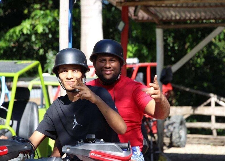 Two people on a motorcycle with helmets, a red shirt, and a black shirt, with a green vehicle and a wooden structure in the background. 3