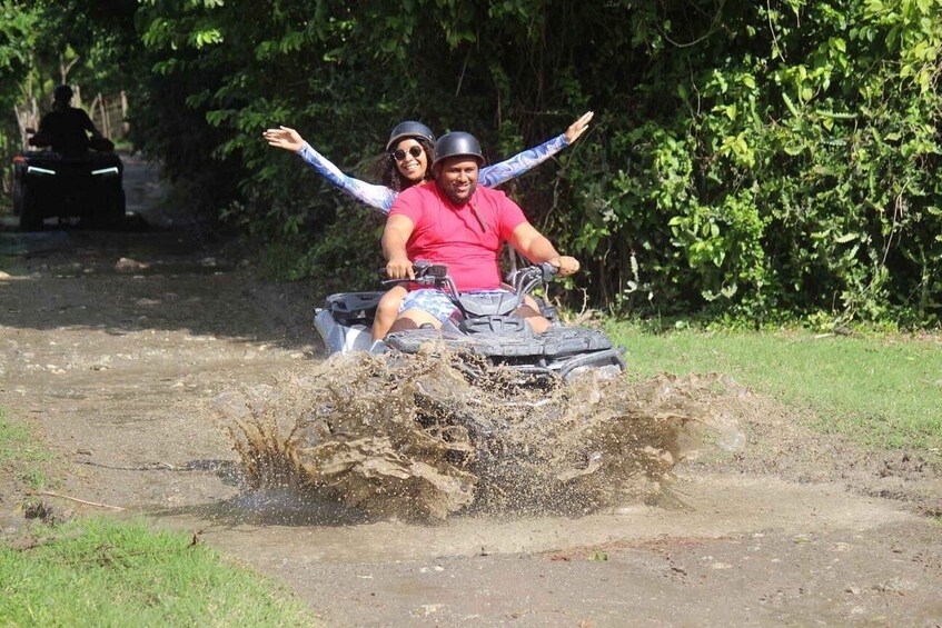 Two people on an ATV in a muddy area with trees in the background. 5
