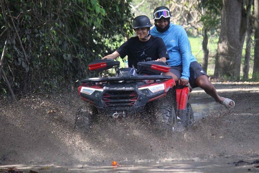 Two people riding a red ATV on a dirt path surrounded by trees. 1