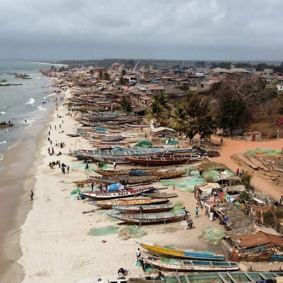 A beach lined with numerous boats, some with people around them, and a town in the background. 1