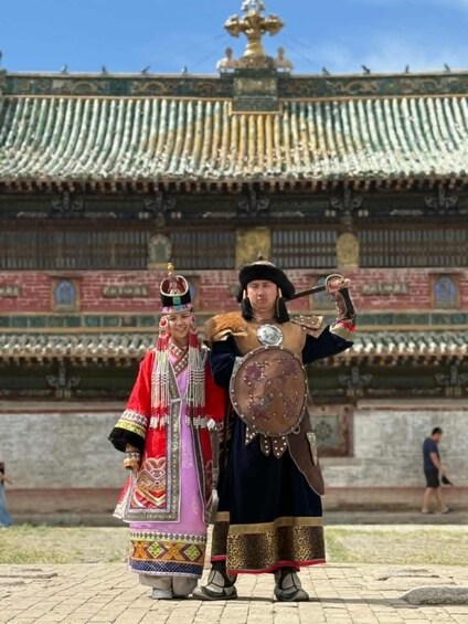 Two individuals in traditional attire standing in front of a historical building with ornate architecture. 1