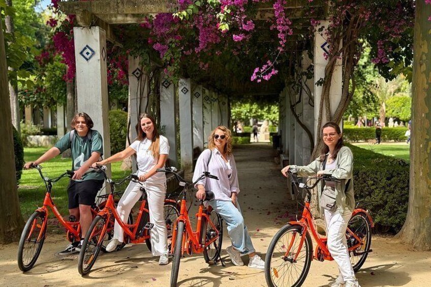 Four people on bicycles in a park with a covered walkway and flowering trees. 3