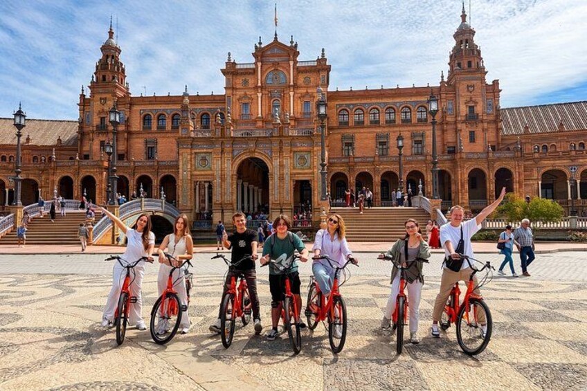 A group of people on bicycles in front of a historic building with arches and towers. 1