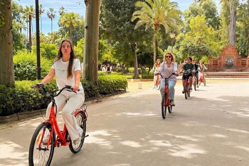 A group of people riding bicycles on a paved path in a park with trees and a monument in the background. 2