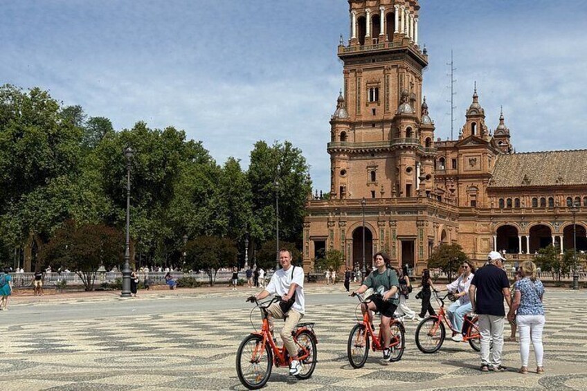A plaza with people on bicycles, a large historic building with a bell tower, and trees in the background. 4
